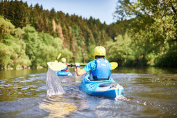 Ein Kajakfahrer ist von hinten zu sehen, wie er durch klares Wasser paddelt. Der Fluss schlängelt sich durch grüne Landschaft des Bayerischen Waldes.