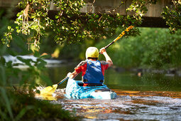 Kajakfahrer mit gelbem Helm paddelt unter einer Brücke auf einem Fluss im Bayerischen Wald.